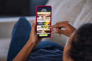 Woman lying on couch using food delivery app on smartphone. Close up of african woman hands holding cellphone and ordering food online. Girl using smart phone to get home delivered take away lunch.